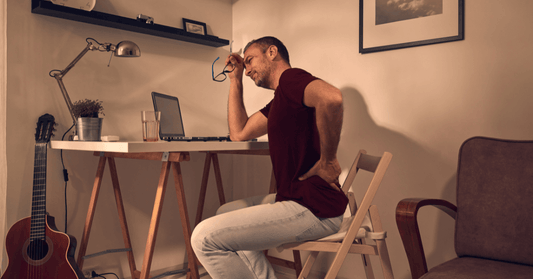 Man sitting at a home office desk clutching his lower back in pain, indicative of poor ergonomic posture potentially leading to sciatica discomfort.